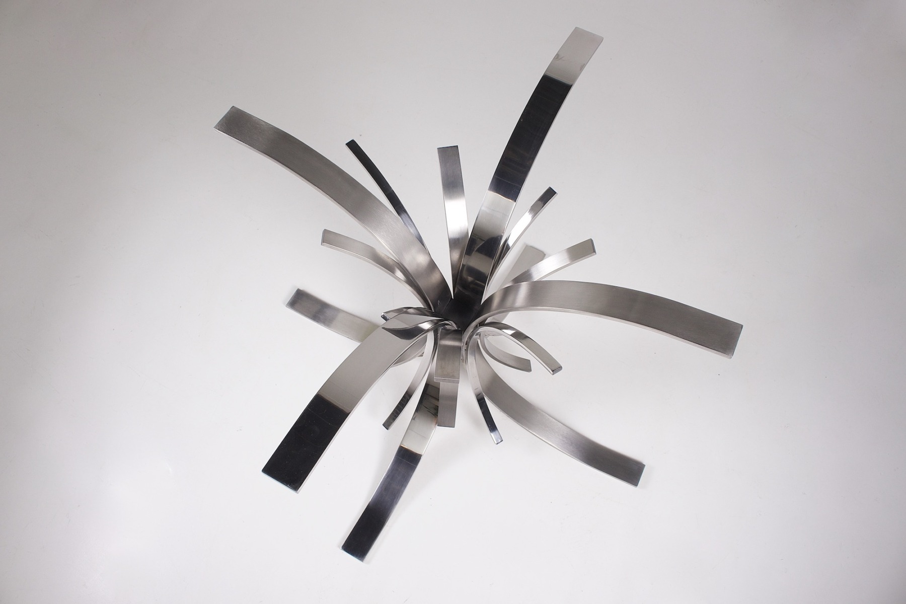 Des bandes métalliques incurvées rayonnent à partir d'un point central, faisant écho à la forme dynamique de la table « Gerbe 56 A » de Maria Pergay, pour former une sculpture abstraite en forme d'étoile sur un fond blanc uni.