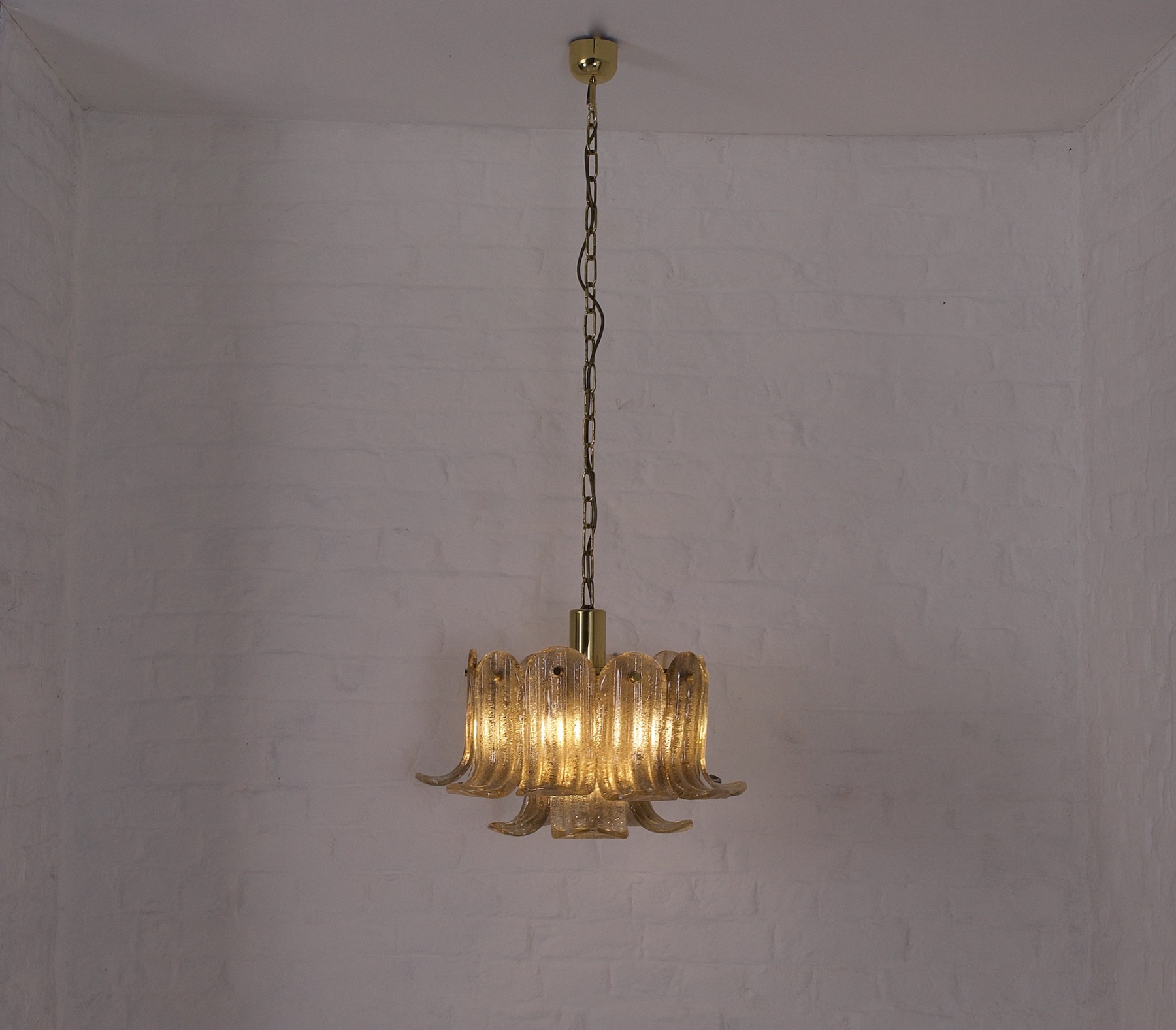 Une suspension en verre texturé aux nuances ambrées, composée de panneaux ondulés délicatement façonnés en forme de pétales. Suspendue à une chaîne dorée assortie à sa structure centrale en métal, elle diffuse une lumière chaleureuse et enveloppante, idéale pour créer une ambiance élégante